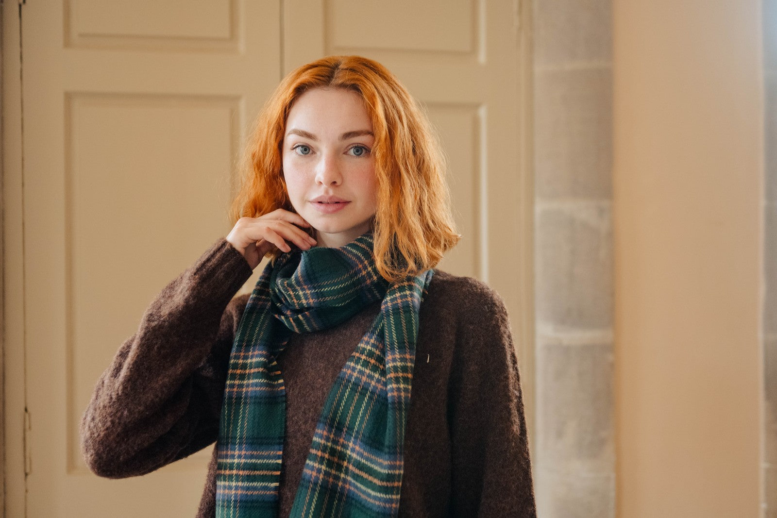 Woman wearing a plaid scarf and brown coat indoors.