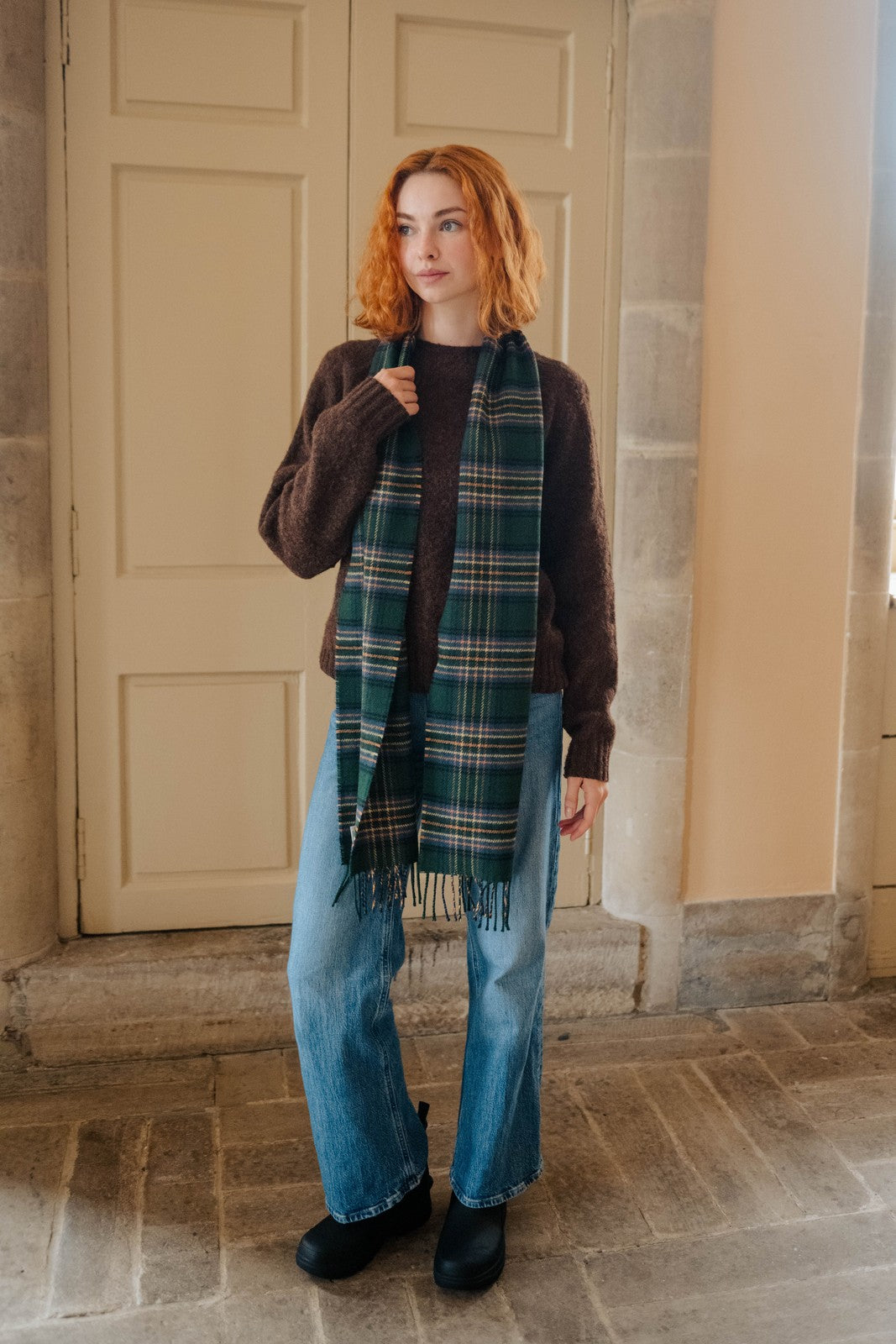 Woman wearing a plaid scarf and brown sweater in front of a stone building.