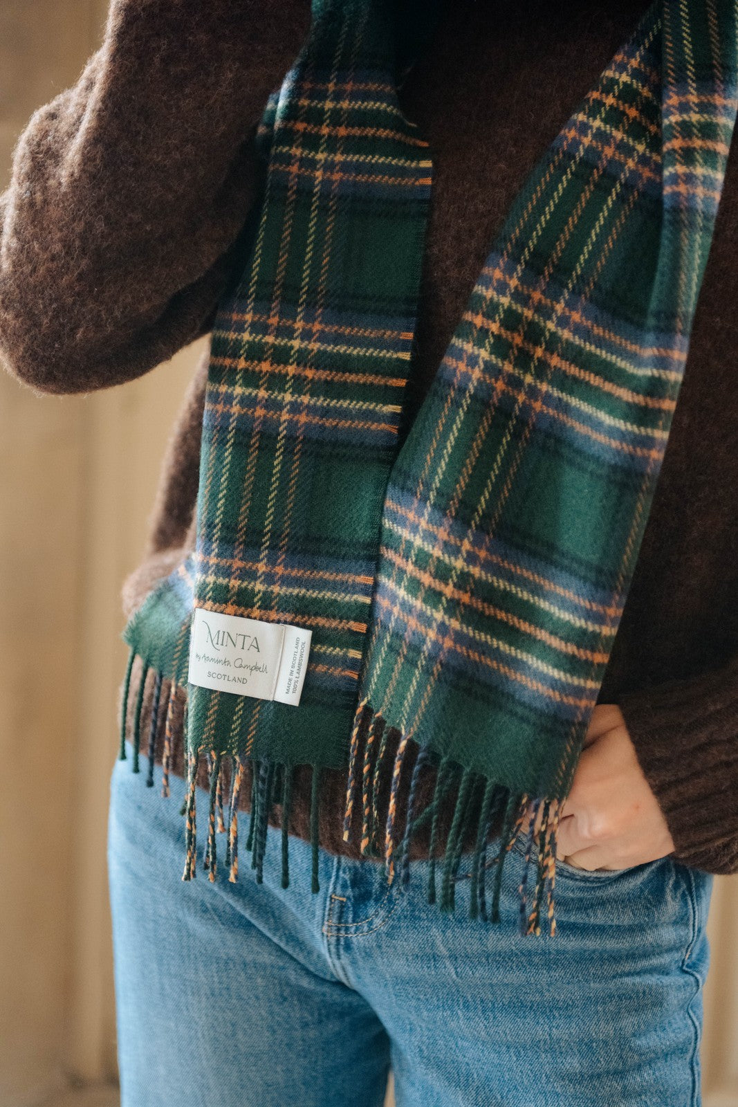 Person wearing a green plaid scarf with a visible brand label, standing indoors.