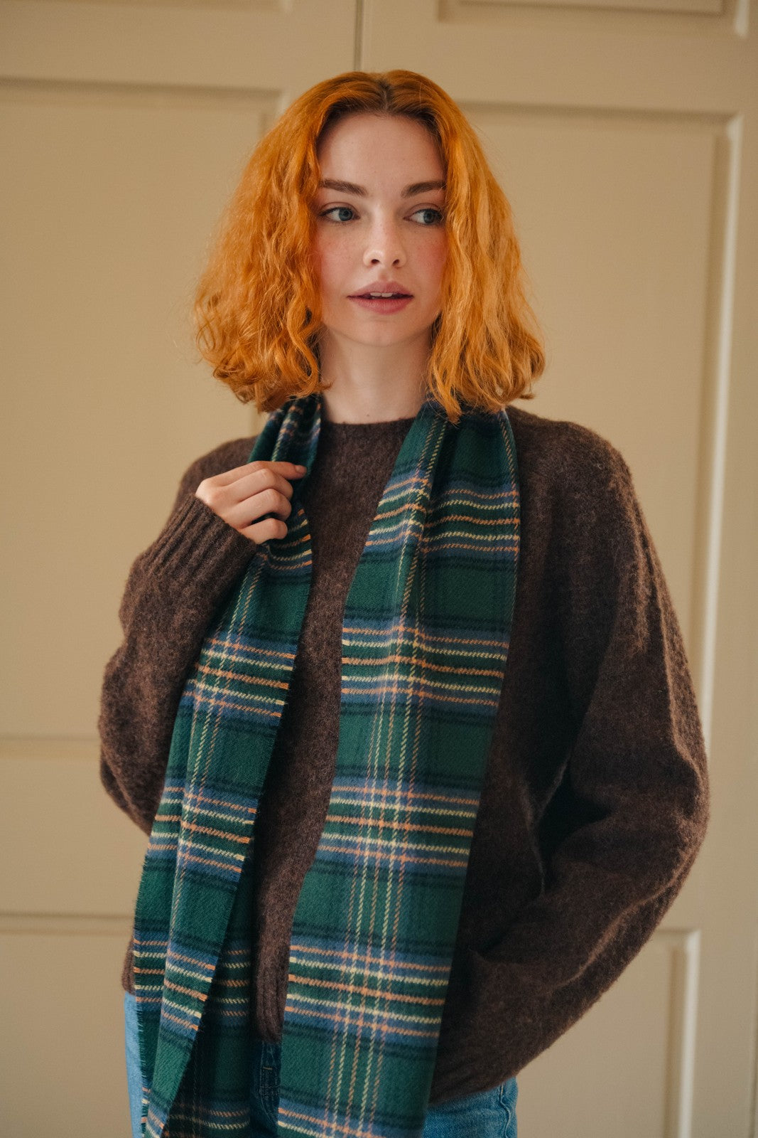 Woman wearing a brown sweater and green plaid scarf against a beige wall.