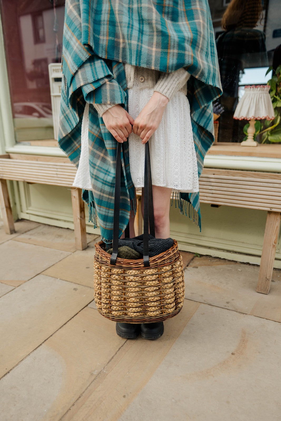 Person holding a woven basket with shoes, wearing a plaid shawl and white dress.