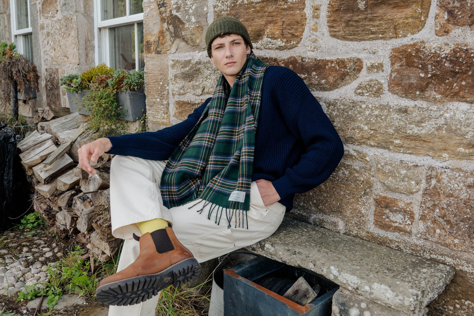 Person wearing a plaid scarf and dark jacket sitting against a stone wall.