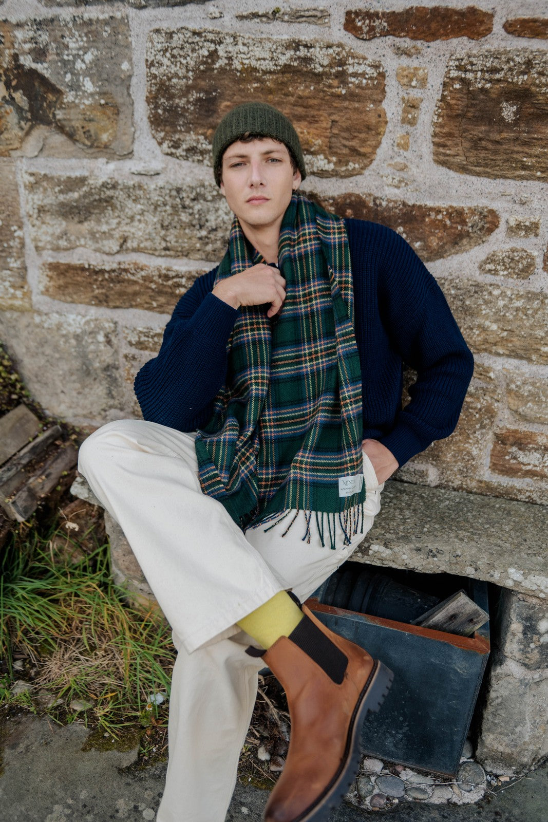 Person wearing a navy sweater, plaid scarf, and white pants sitting against a stone wall.