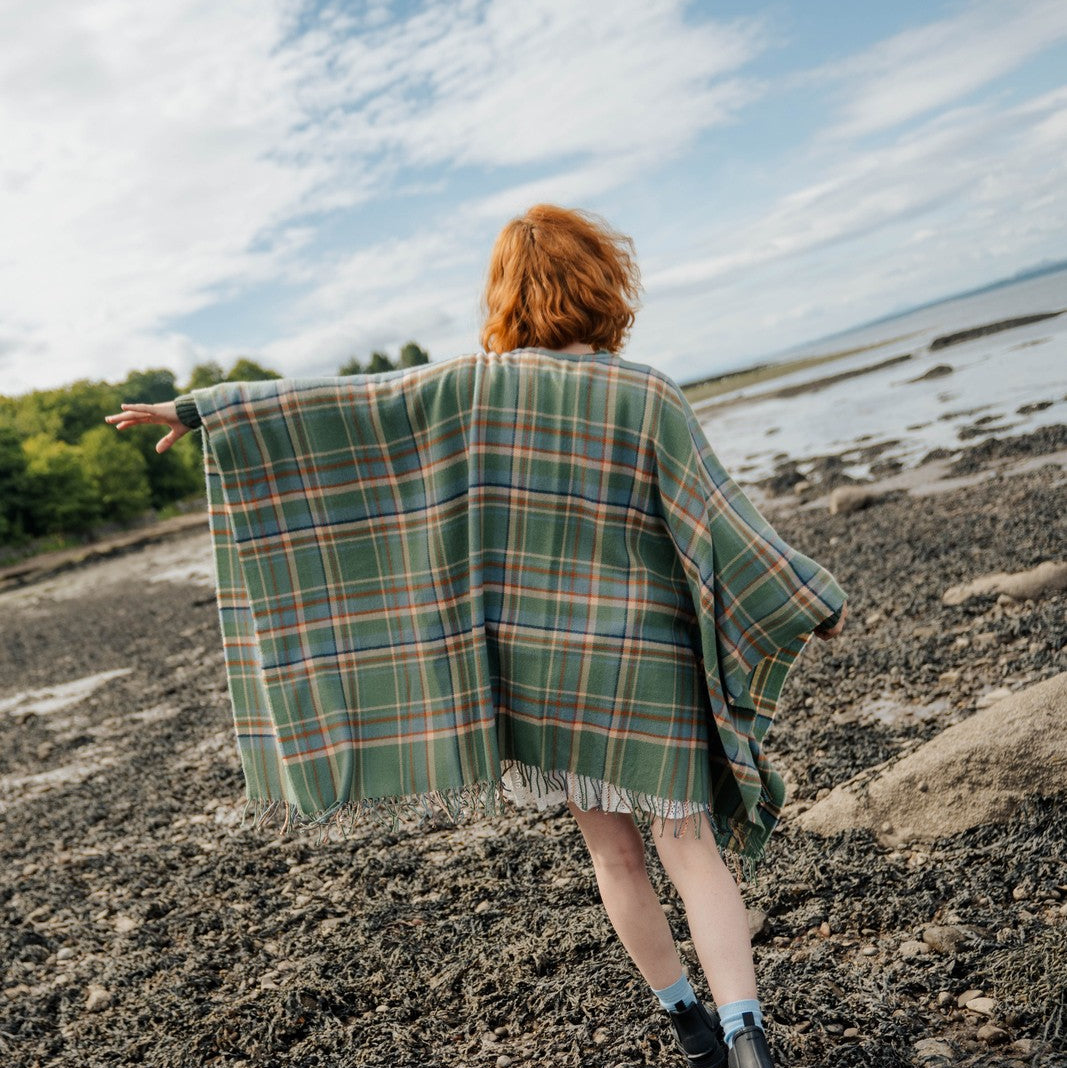 Person with red hair wearing a green plaid blanket over shoulders on a rocky beach.