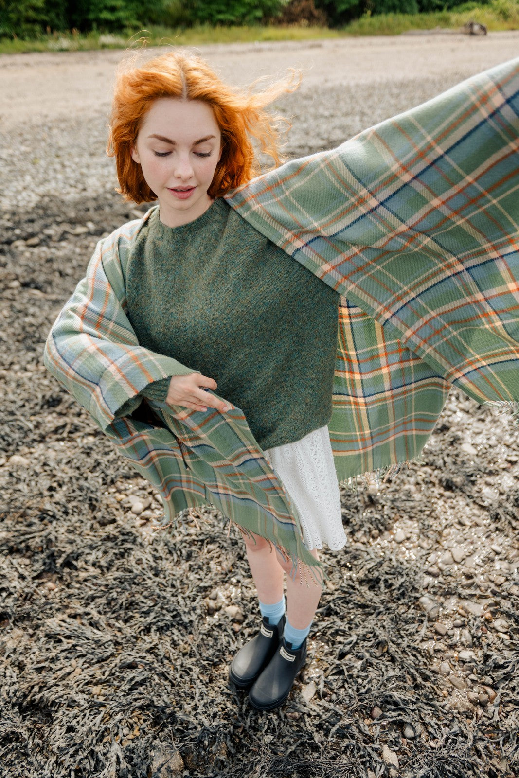 Woman with red hair holding a green plaid blanket outdoors on a gravel path.