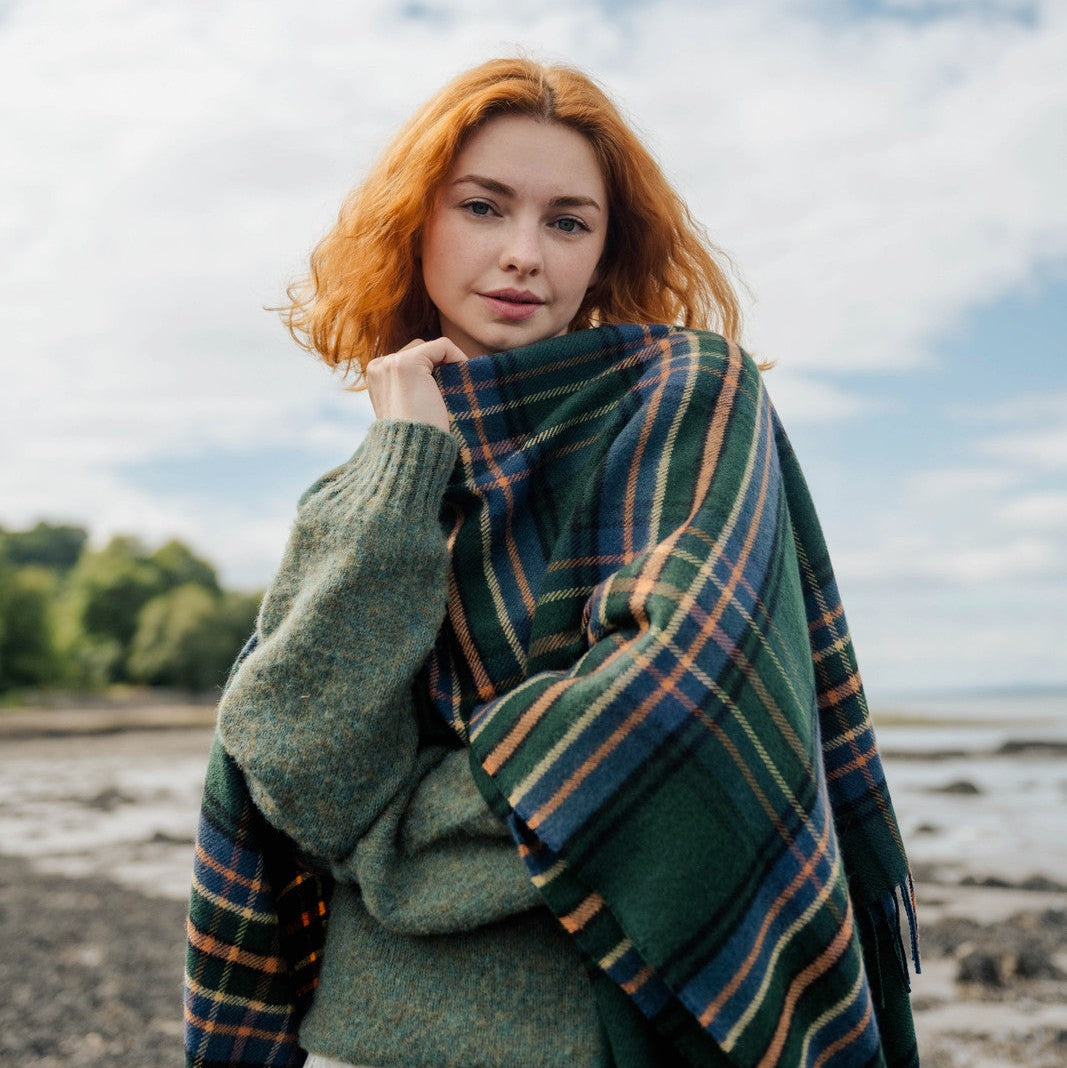 Woman with red hair wrapped in a plaid blanket standing on a beach.