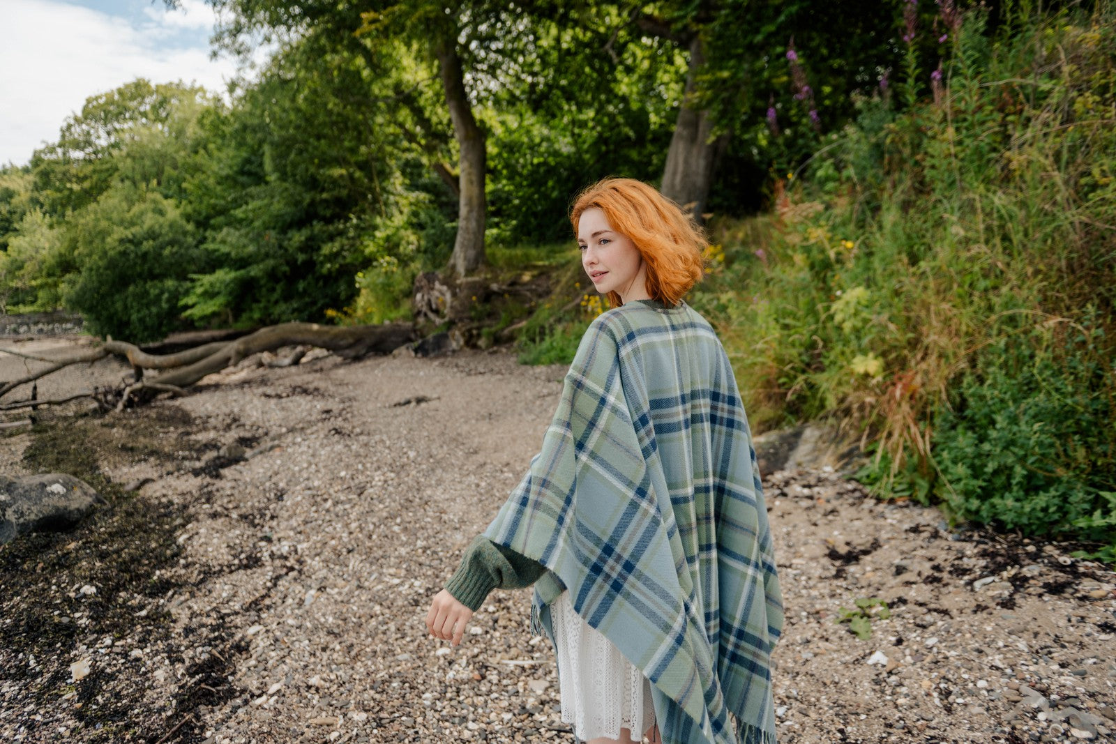 Woman with red hair wearing a green plaid poncho standing in a natural setting with trees and plants.