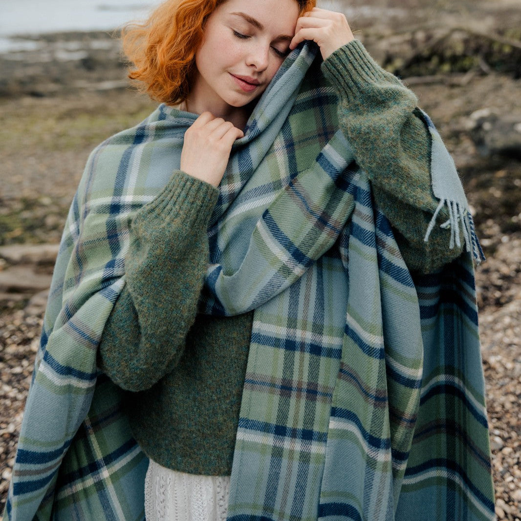 Woman wrapped in a green plaid blanket standing on a rocky beach with trees and water in the background.