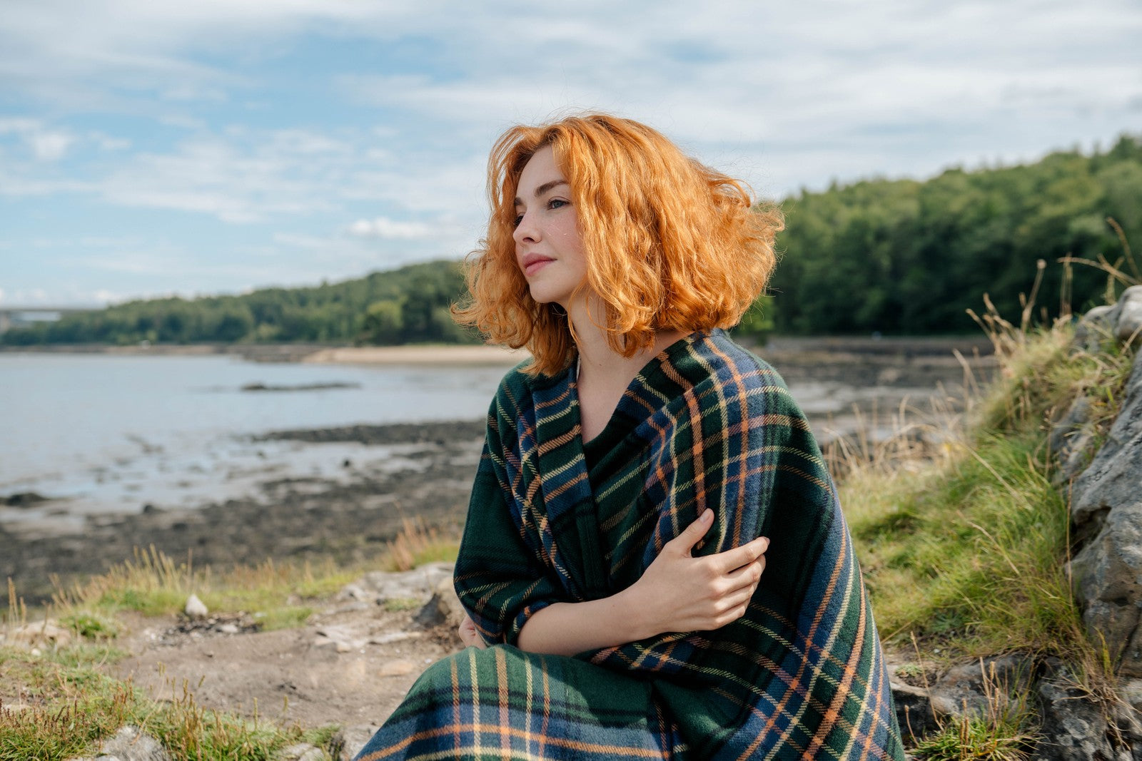 Woman with red hair sitting by a lakeside wearing a plaid coat.