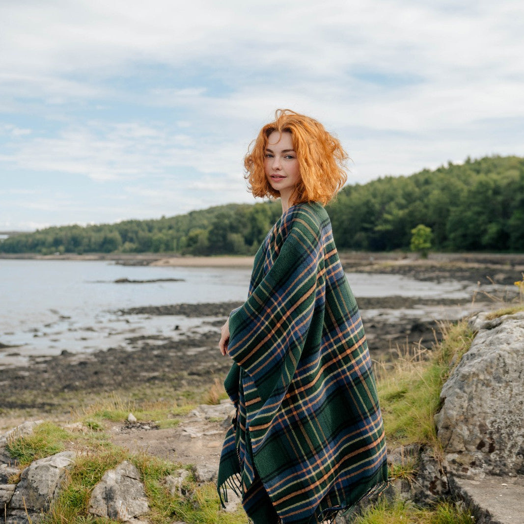 Person with red hair wrapped in a green plaid blanket standing by a body of water with trees in the background.