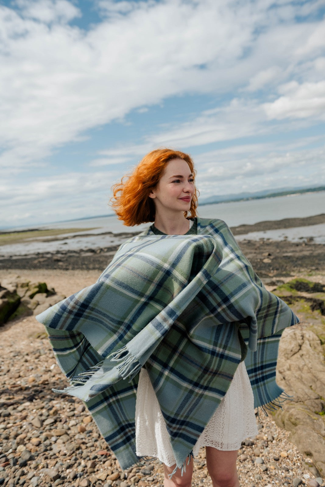 Woman with red hair wearing a green plaid blanket on a rocky beach.