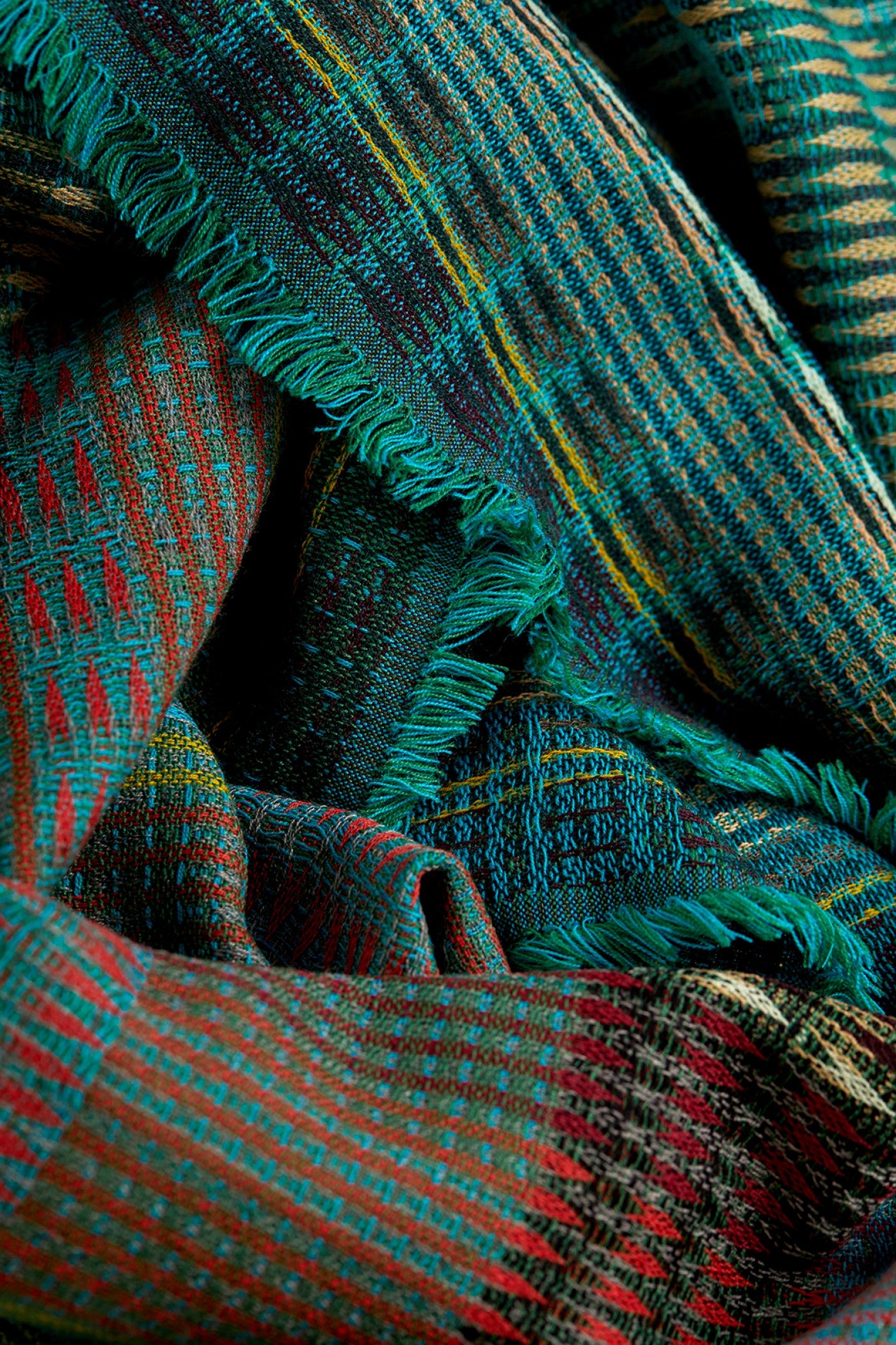 A close-up of a woven plaid scarf with fringed edges, featuring teal green, red, black, and yellow stripes.
