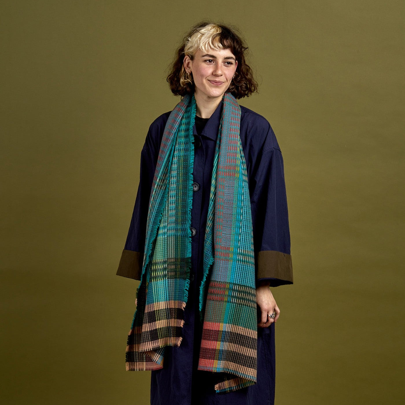 A model stands against a solid olive green studio background wearing a long navy blue coat with brown cuffs, brown trousers, and a teal and red plaid scarf.