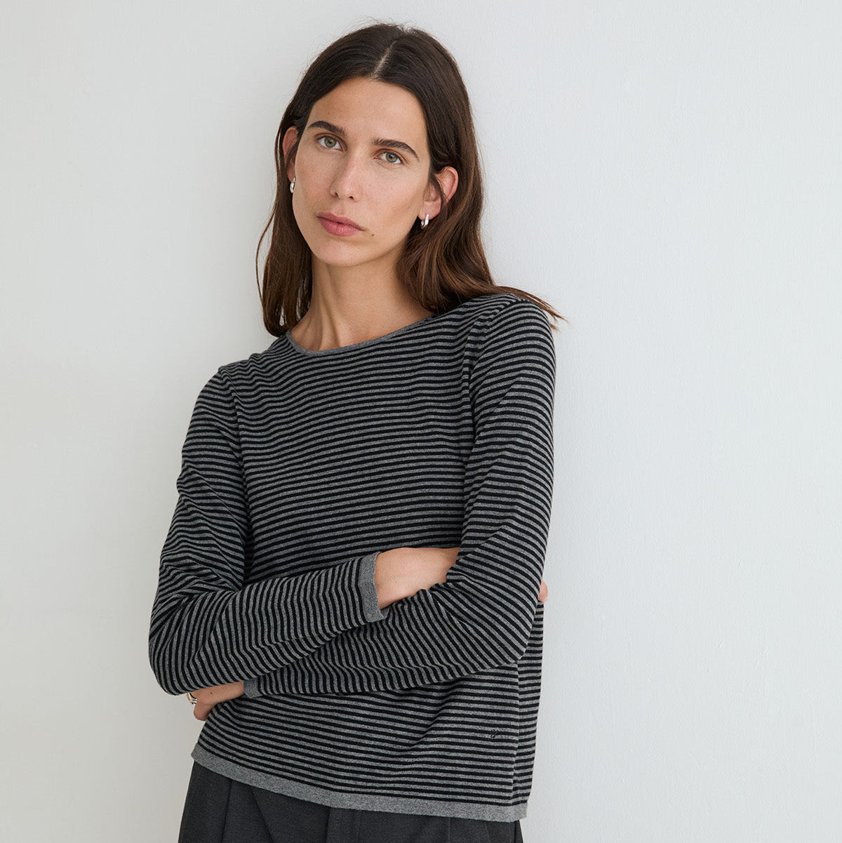 A woman with dark brown hair is wearing a long-sleeve knit top with thin black and gray horizontal stripes, paired with dark gray trousers, standing against a plain light gray wall.