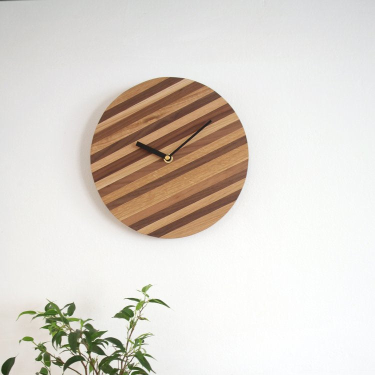 Round wooden wall clock with diagonal light and dark wood stripes, black hands, and gold center.