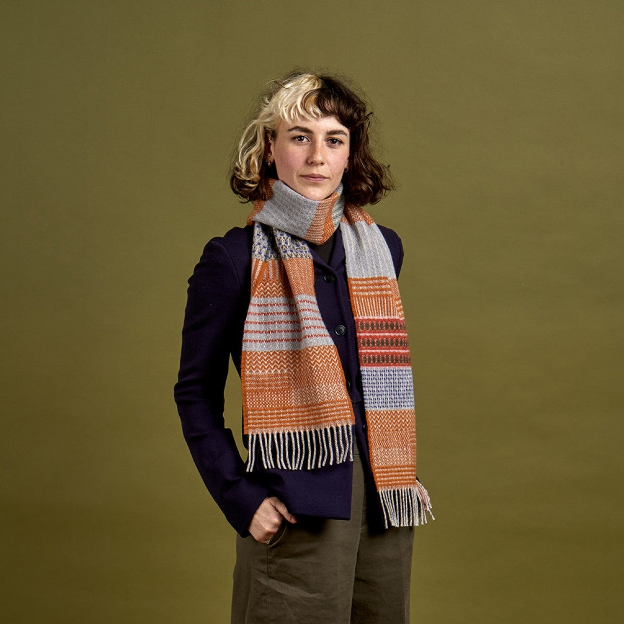 A model stands against a solid olive green studio background wearing a navy cardigan, wide-leg brown trousers, and a thick striped scarf in orange, beige, and blue.