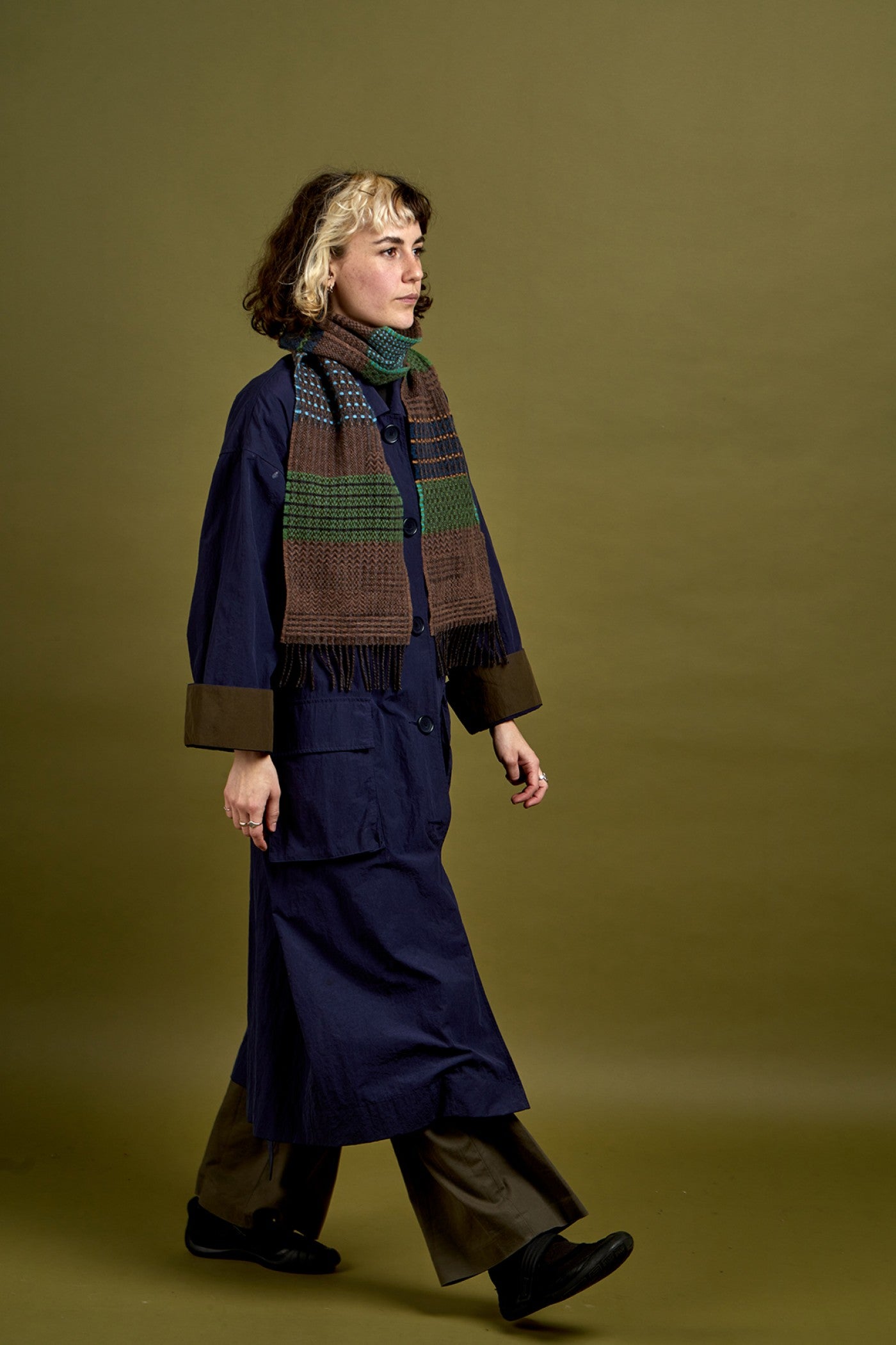 A model walks in profile wearing a dark navy blue coat with brown cuffs, brown trousers, and a thick striped knit scarf in brown, green, and blue, set against a solid olive green studio background.