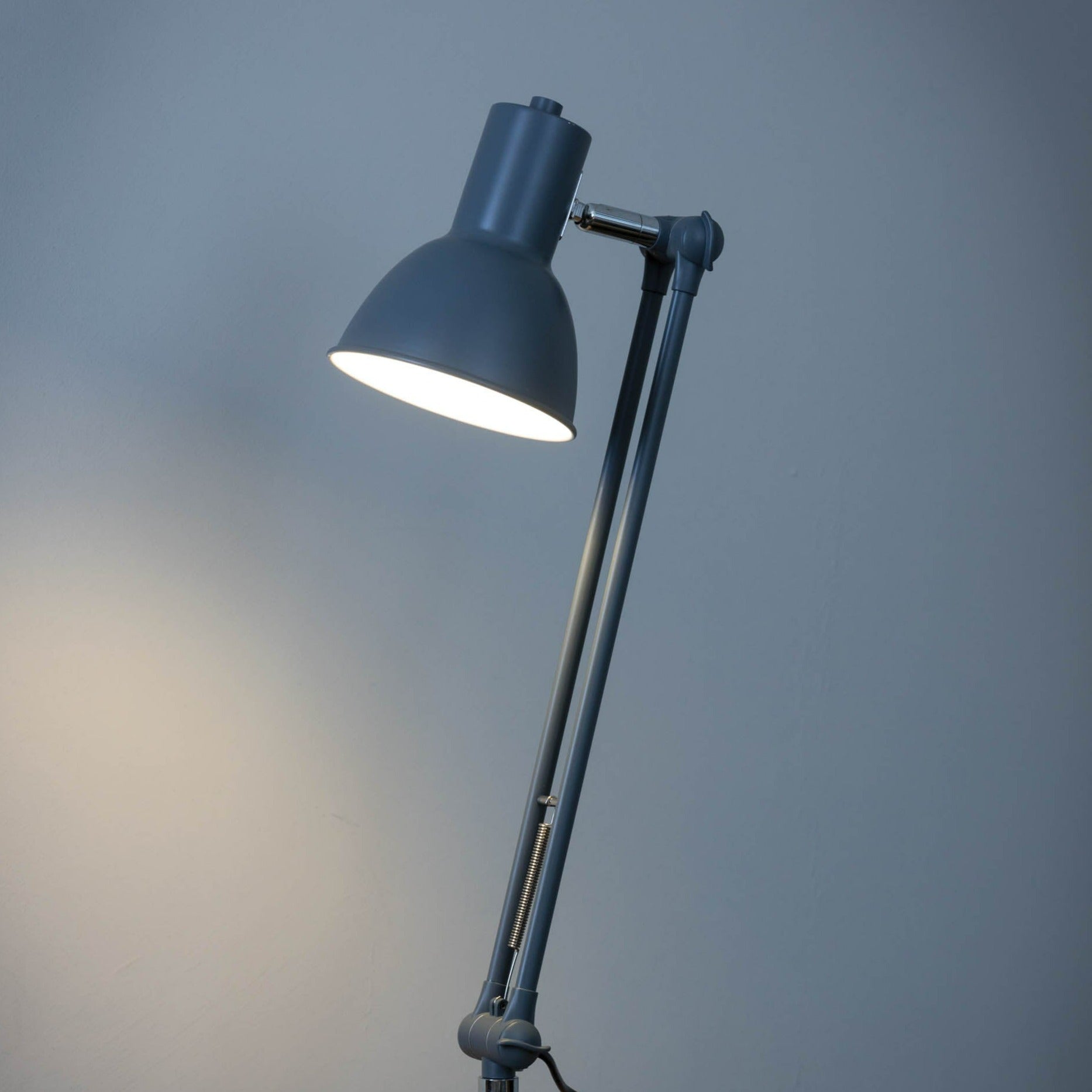 A gray adjustable desk lamp with a dome-shaped shade is illuminated against a light gray wall.