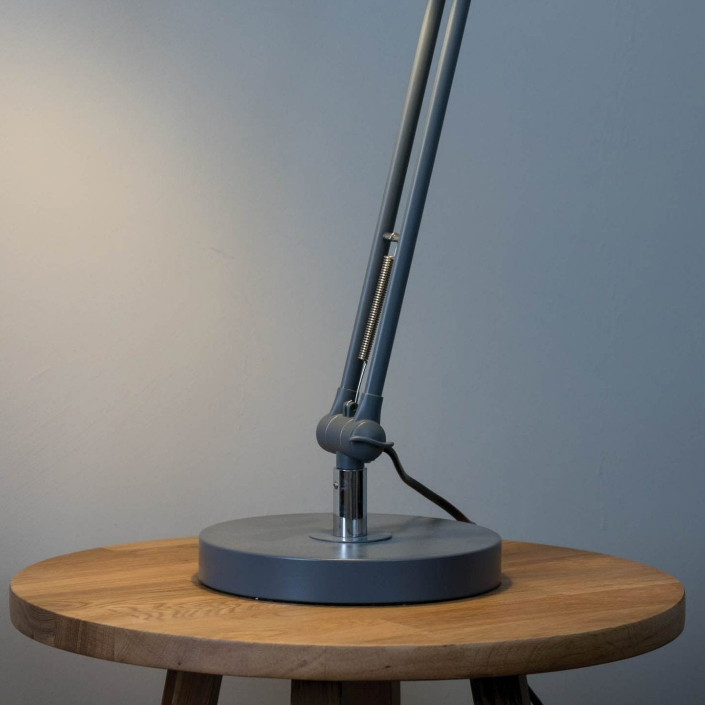 Gray adjustable desk lamp with a circular base, sitting on a light brown wooden table.