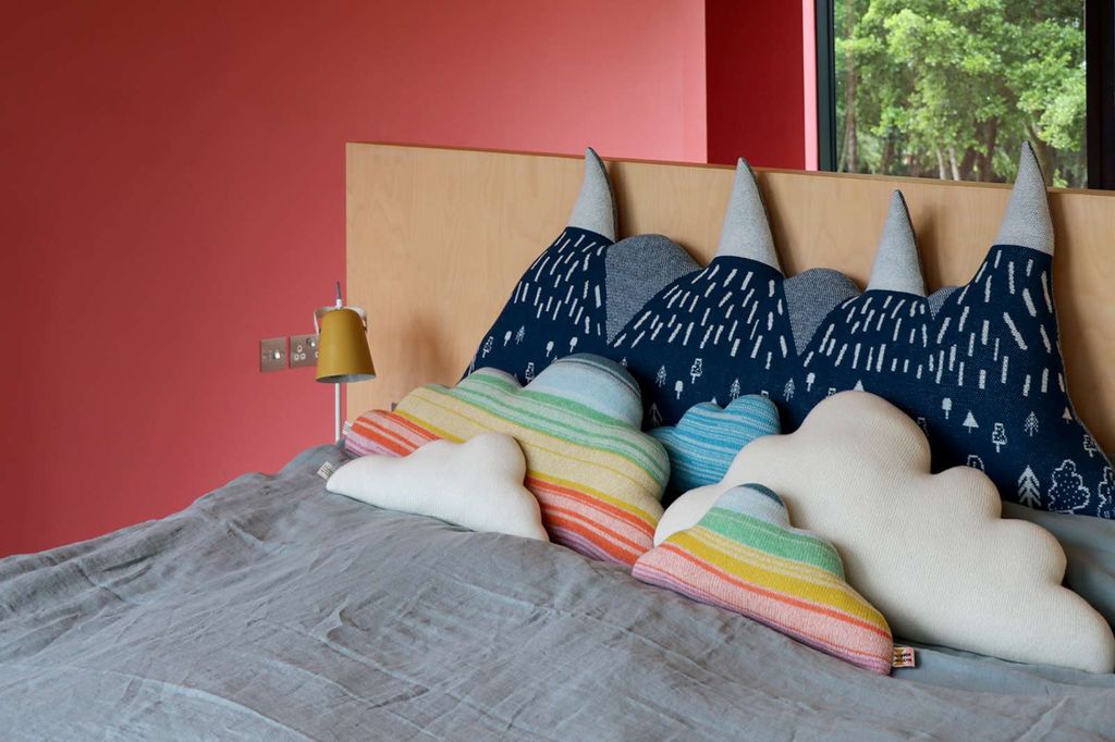 A cozy bedroom scene with a bed made up in gray bedding and decorated with colorful cloud-shaped pillows and dark blue mountain-patterned pillows against a light wood headboard. The wall is a muted coral-red color, and a window in the background shows green trees.