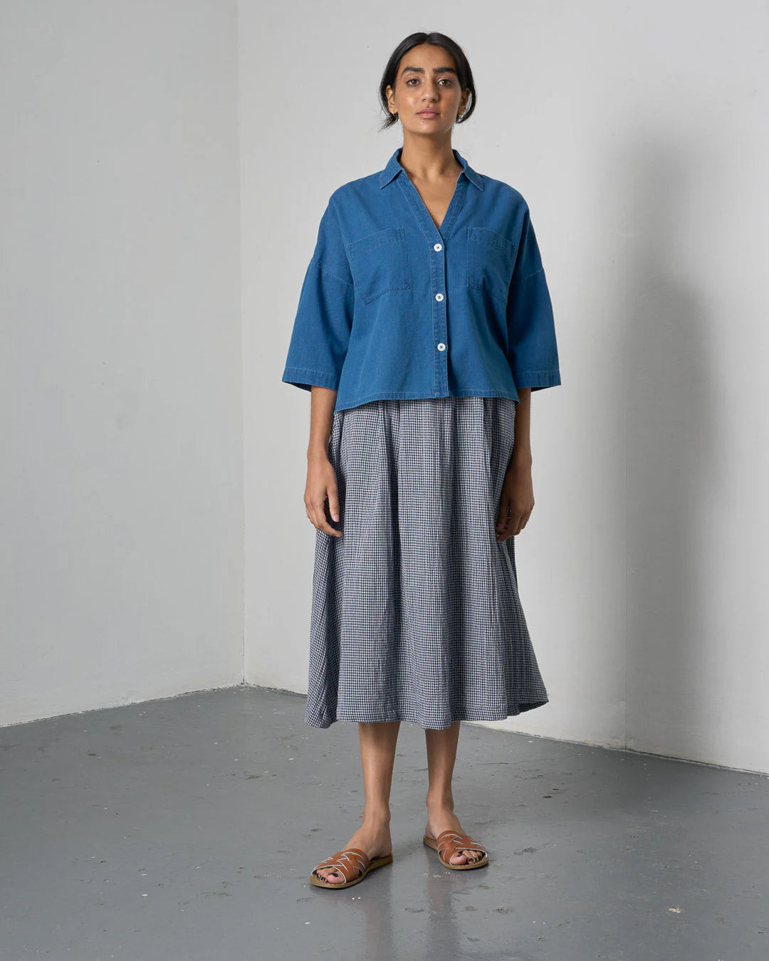Woman wearing a blue shirt and gray skirt standing against a white wall.