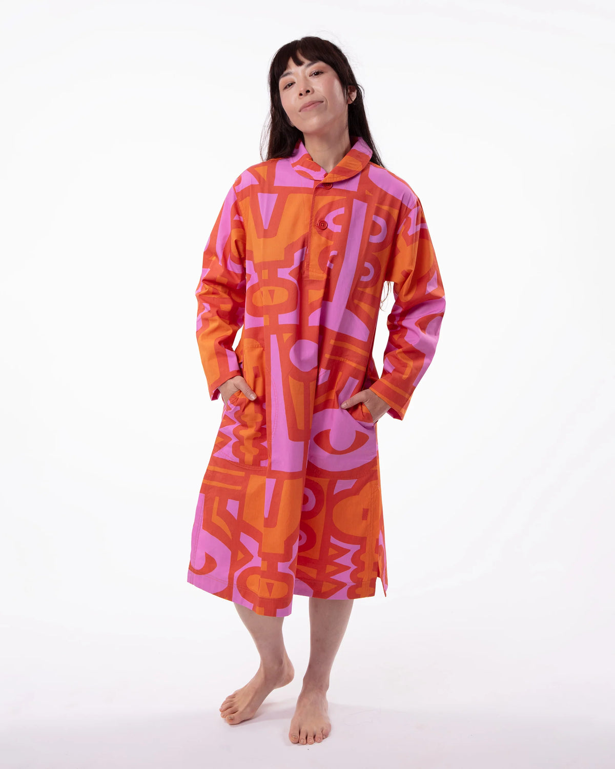 A woman modeling a long-sleeved hooded robe with a bold abstract geometric pattern in orange and pink, standing against a plain white studio background.