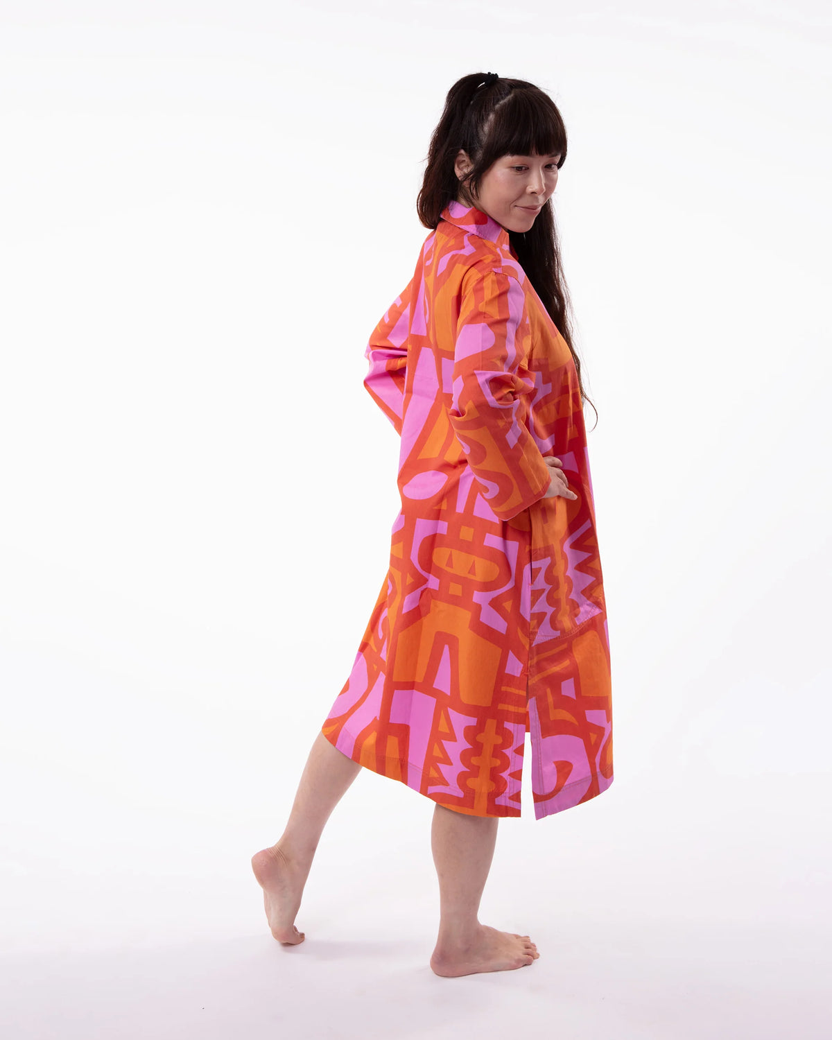 A woman wearing a vibrant orange and pink abstract patterned kimono robe, captured from a side-back angle against a clean white studio background.