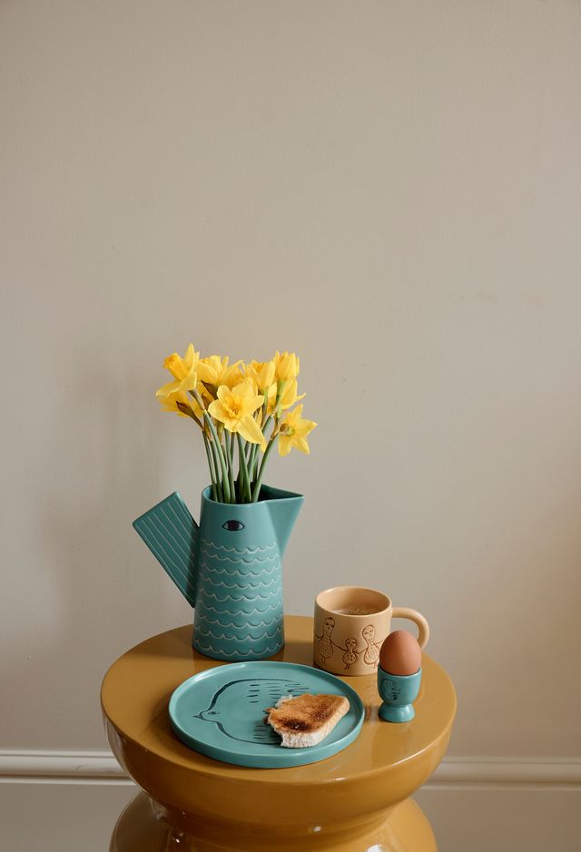A styled tabletop scene with a turquoise pitcher vase holding yellow daffodils, a matching turquoise plate with a piece of toast, a beige patterned mug, and an egg in a turquoise holder, all arranged on a round mustard-yellow pedestal against a plain beige wall.