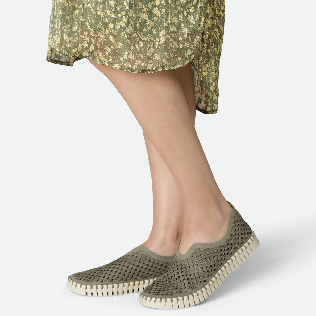 Woman's legs in olive green floral dress wearing olive green perforated slip-on shoes with cream sole.