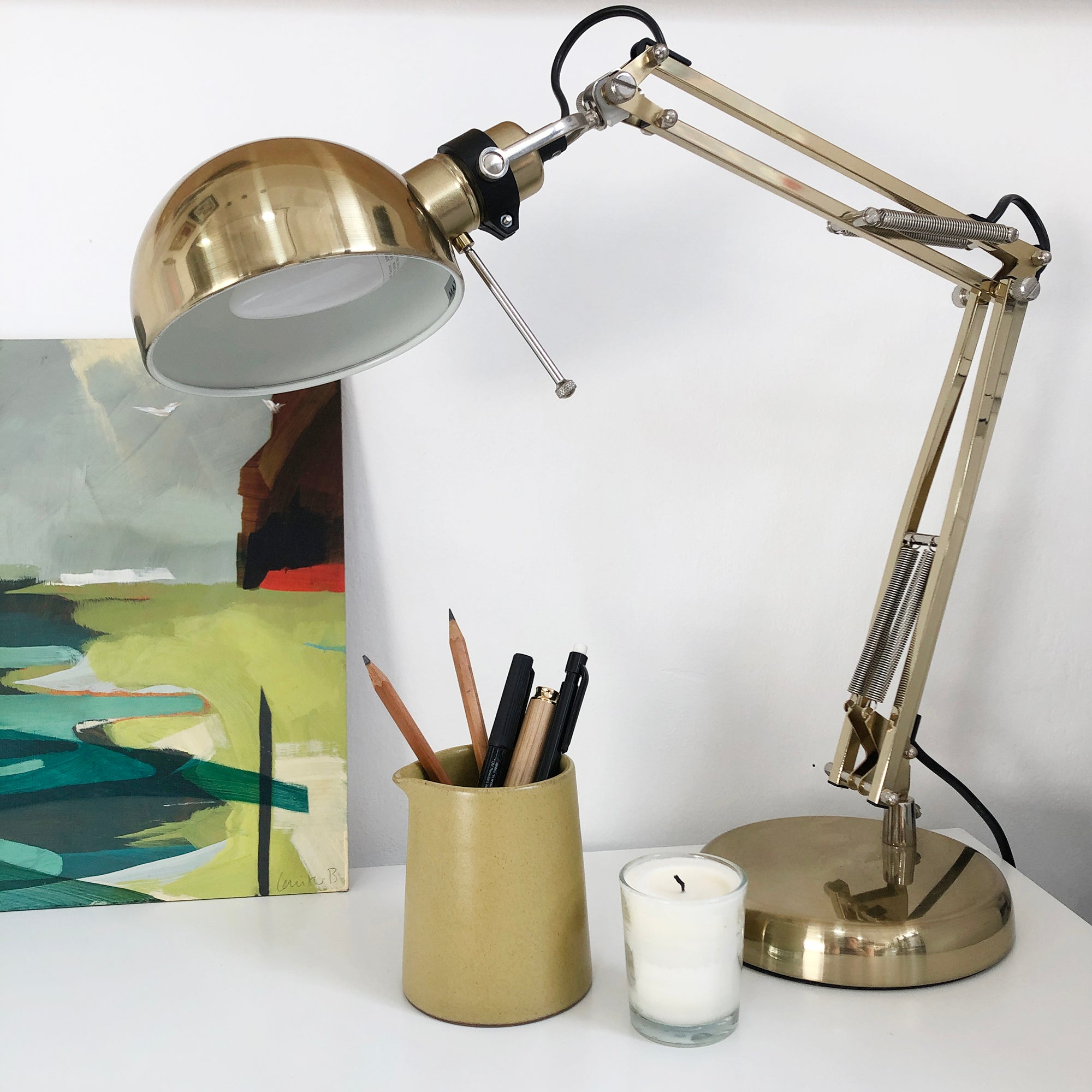 Gold adjustable desk lamp with a dome shade, sitting on a white surface next to a small ceramic holder with writing utensils