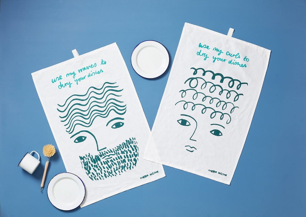 Two white tea towels by Donna Wilson, featuring teal illustrations and text: one with wavy lines, the other with curls.