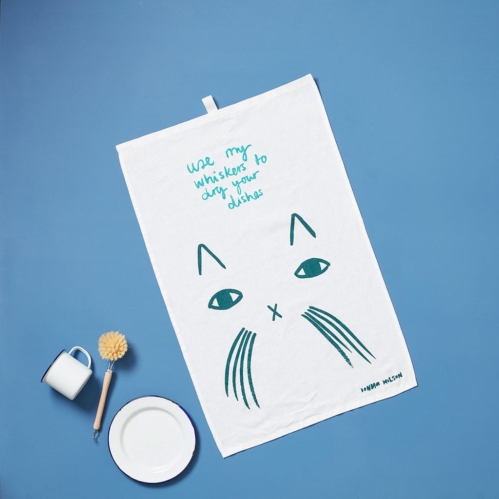 A white tea towel with teal cat design and text: "Use my whiskers to dry your dishes." Brand: Donna Wilson.