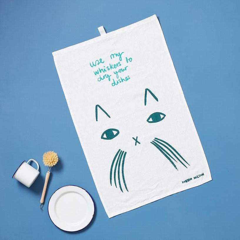 White towel with teal cat design and text, by Donna Wilson, on a blue background with dishes and a scrub brush.
