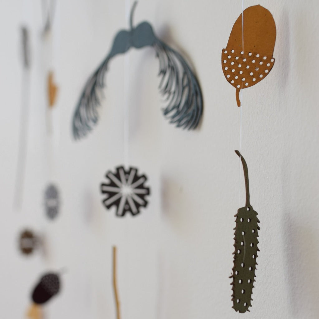 Mobile of laser-cut wood shapes: acorn, seed pods, and geometric designs, suspended from thin threads against a white wall.