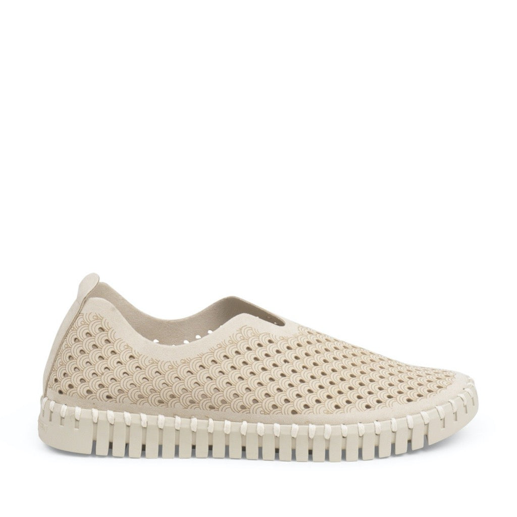 Beige slip-on shoe with a textured, perforated upper featuring a repeating wave pattern. The sole has a unique scalloped edge.