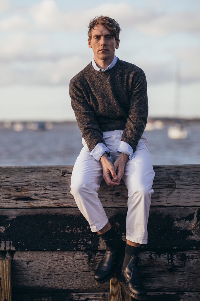 a male model with hands held together, sitting on a wooden bench, wearing a Harley of Scotland Men's Jumper -Staffa, with a background of boats on the sea