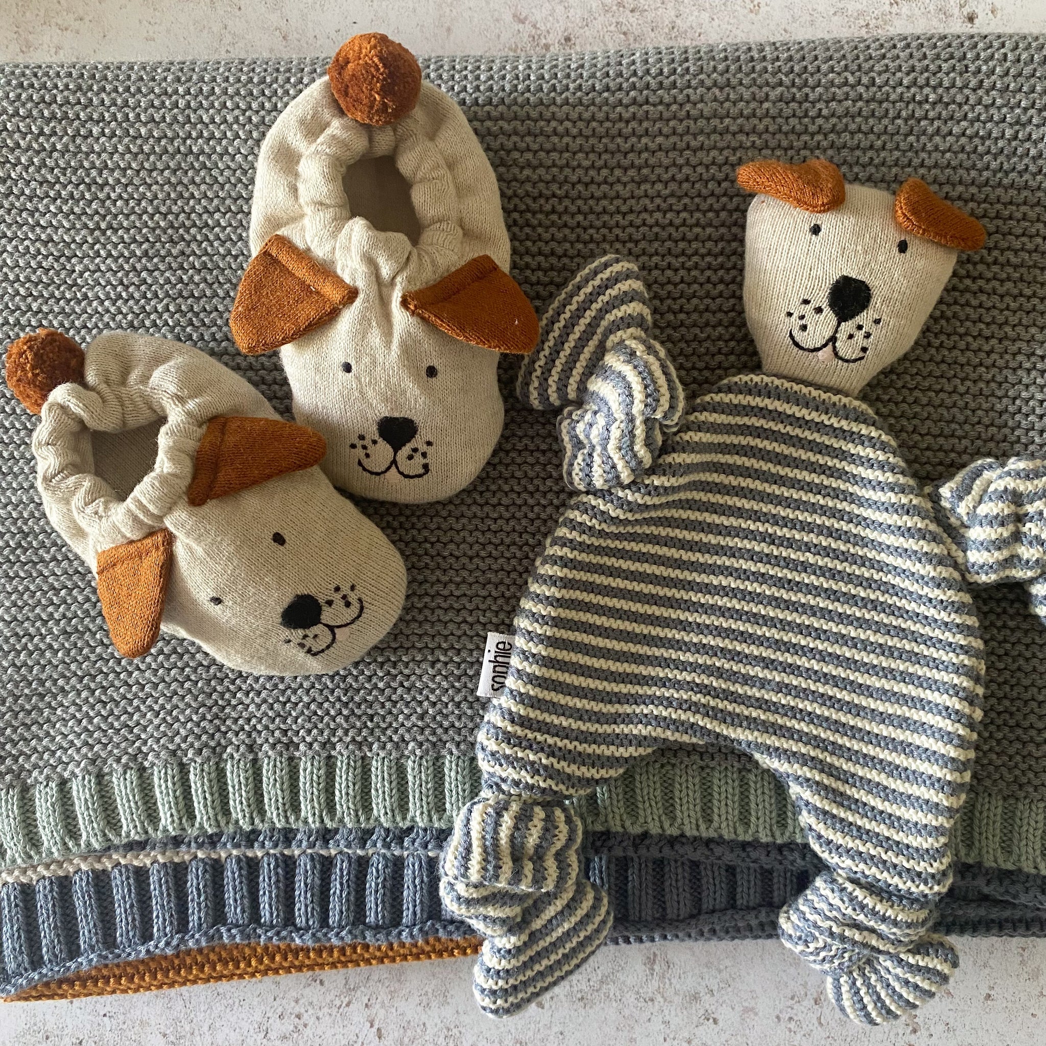 Knitted baby booties and a striped dog-shaped comforter.  The booties and comforter are cream and grey with orange accents.
