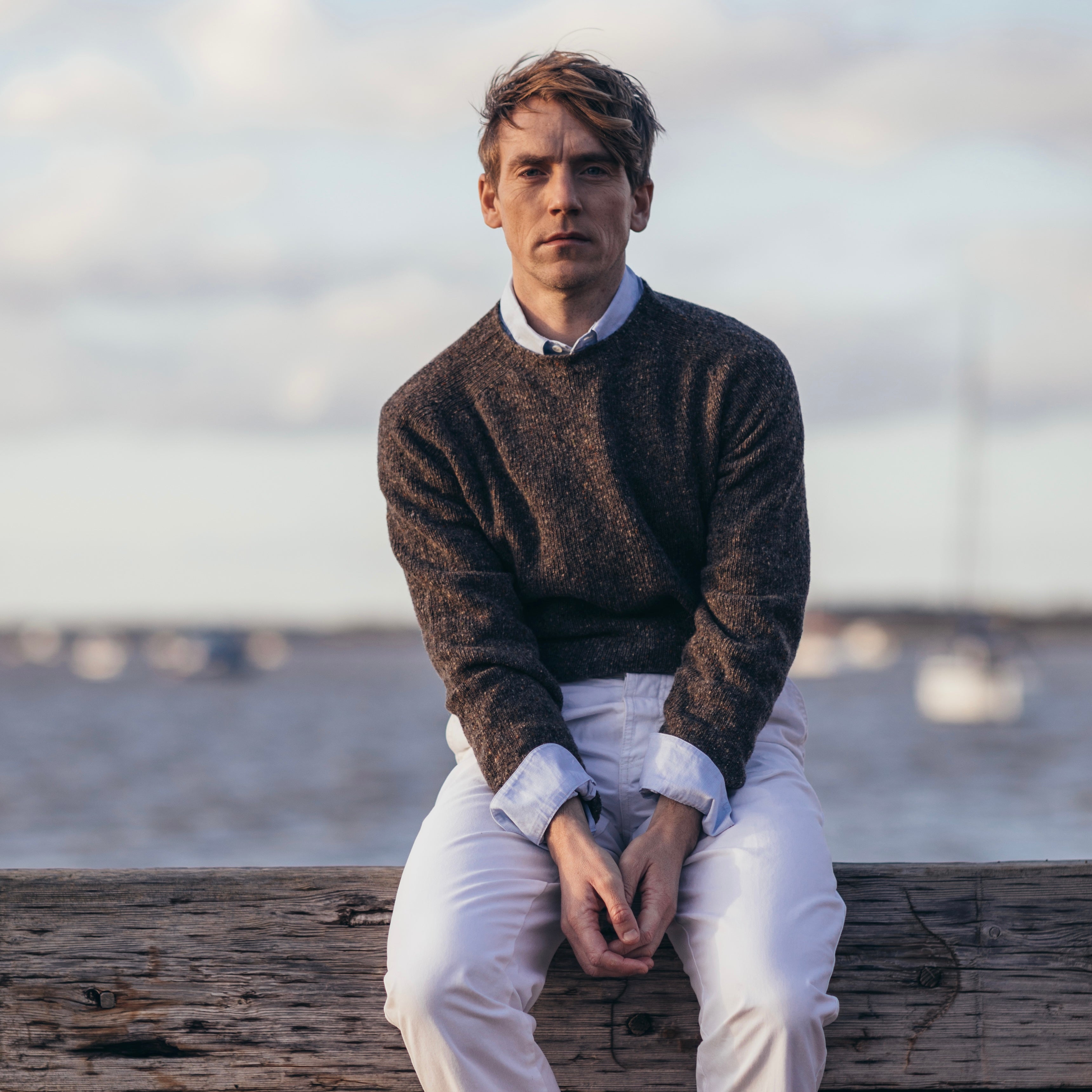 a male model with hands held together, sitting on a wooden bench, wearing a Harley of Scotland Men's Jumper -Staffa, with a background of boats on the sea
