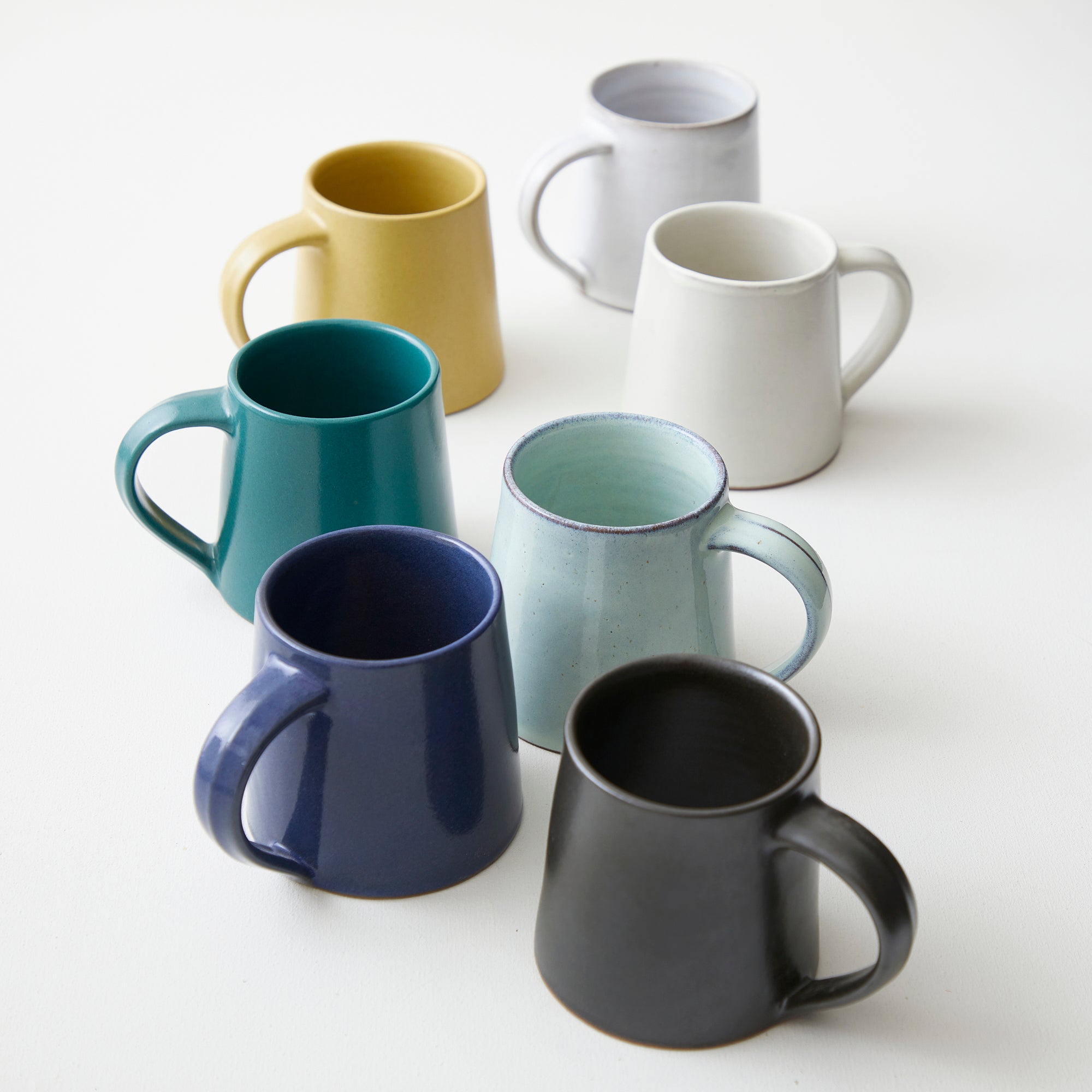 Six ceramic mugs in various colors: mustard yellow, teal, navy blue, off-white, light blue-green, and dark gray.
