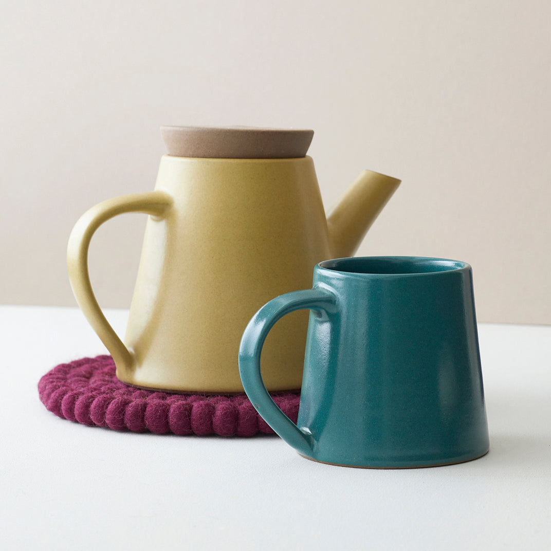 Mustard yellow ceramic teapot with lid and teal ceramic mug on a maroon coaster.