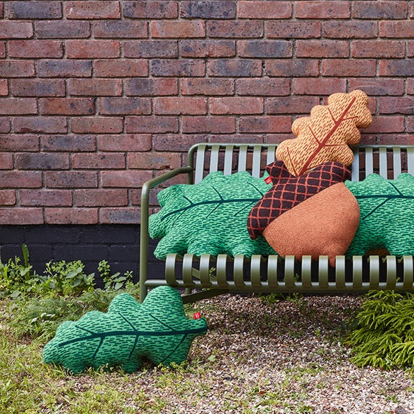 Green leaf and brown acorn cushions by Donna Wilson on a grey bench against a brick wall.