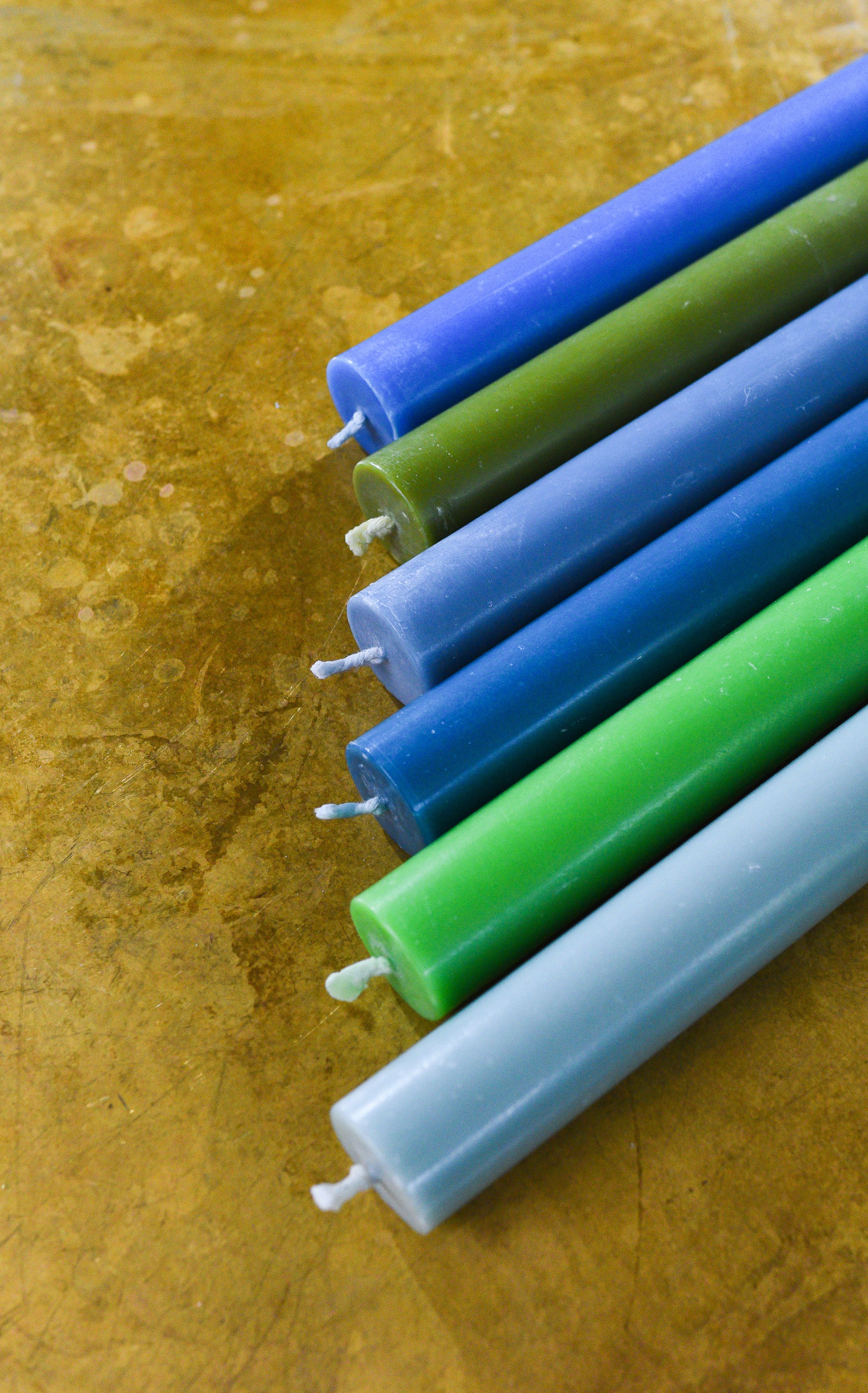 Five long cylindrical candles in varying shades of blue and green, arranged diagonally on a textured gold surface.