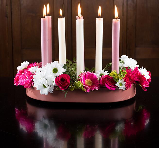 Six tall candles in pink and white, arranged in a mauve oval holder surrounded by a ring of pink and white flowers.