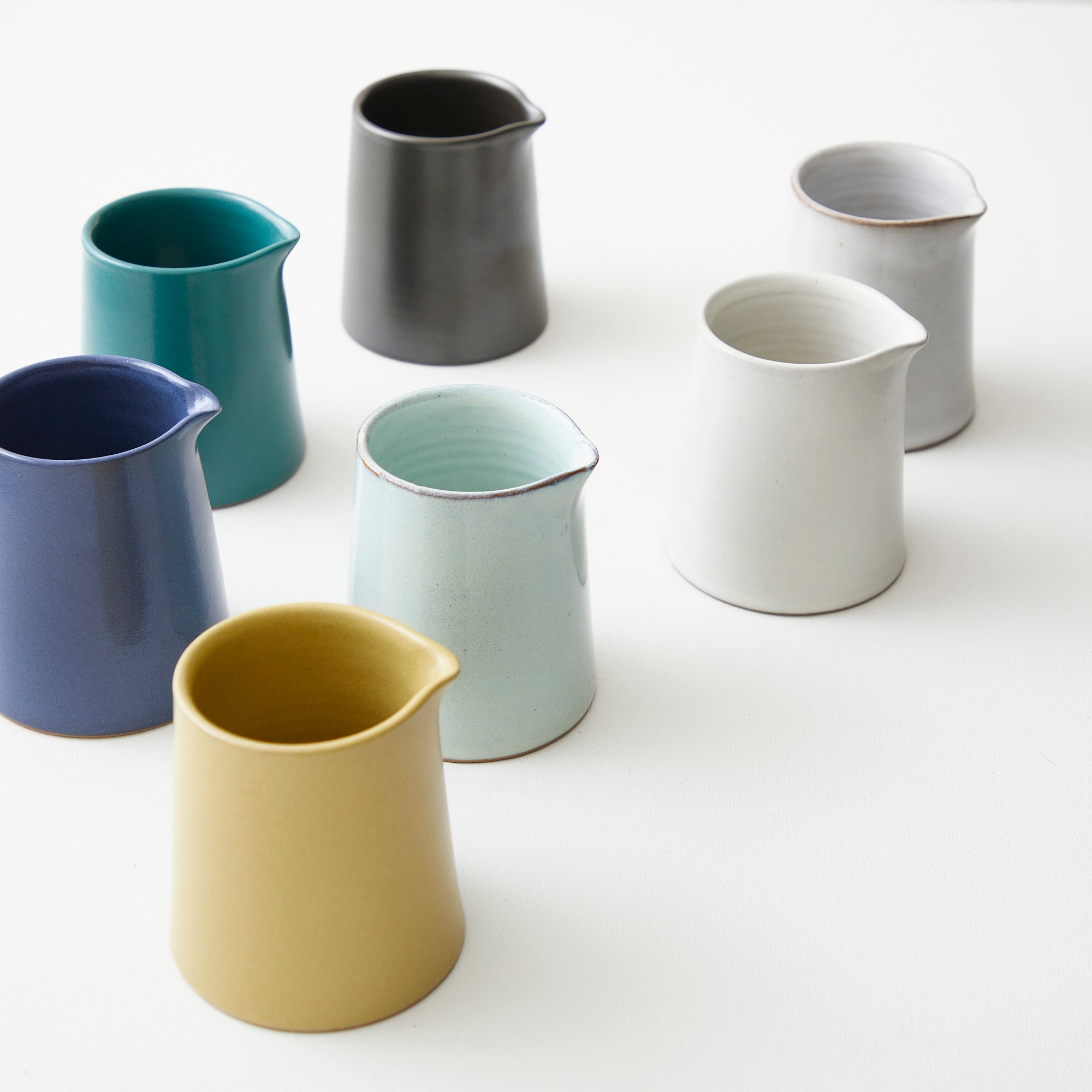 Six small ceramic milk jugs in teal, gray, pale blue, off-white, dark blue, and mustard yellow.