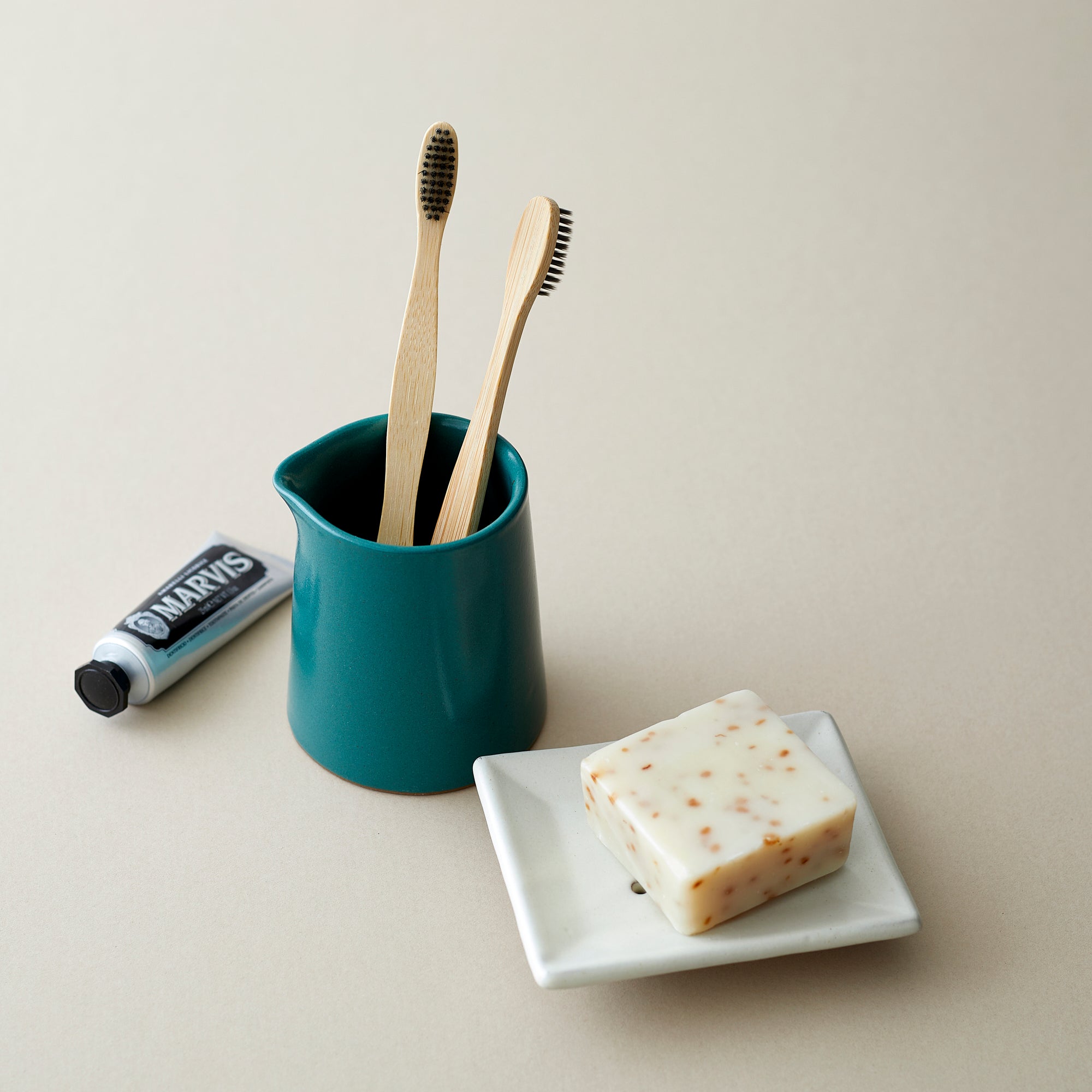 Two bamboo toothbrushes in a teal ceramic holder, next to a bar of speckled soap on a white soap dish.