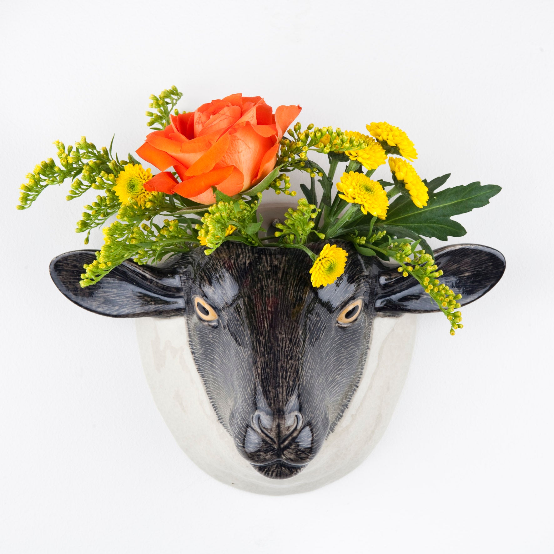 A ceramic wall vase in the shape of a black faced Suffolk sheep, decorated with a colorful floral arrangement.