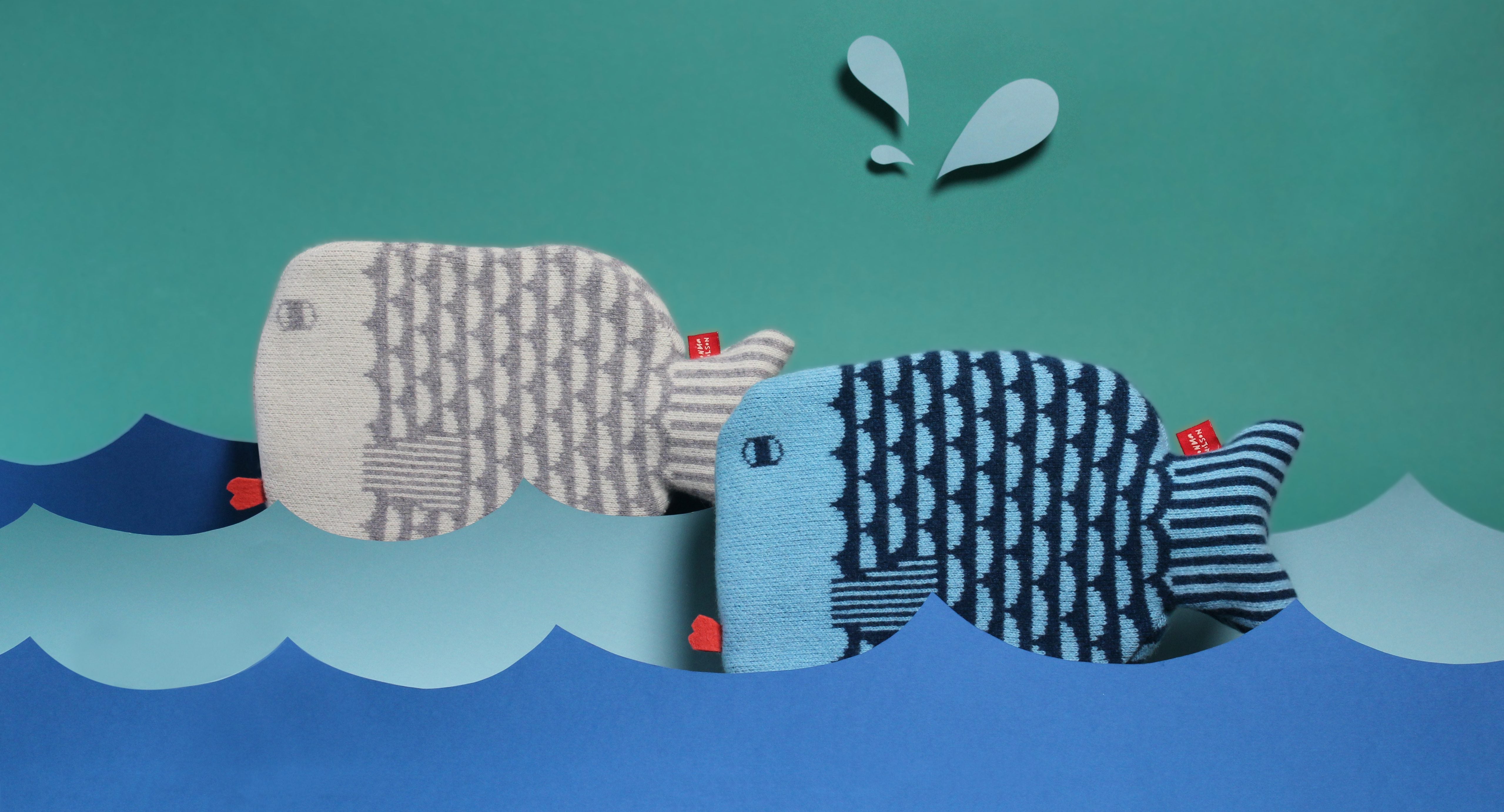 Two plush fish shaped hot water bottles by Donna Wilson, one gray with white scales, the other blue with dark blue stripes, in a colorful sea.