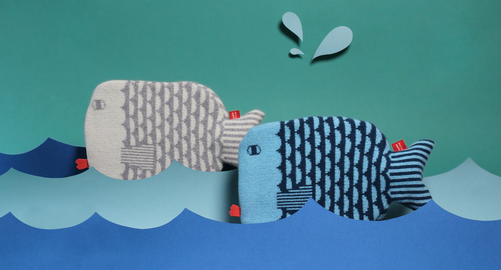 Two plush fish shaped hot water bottles by Donna Wilson, one gray with white scales, the other blue with dark blue stripes, in a colorful sea.