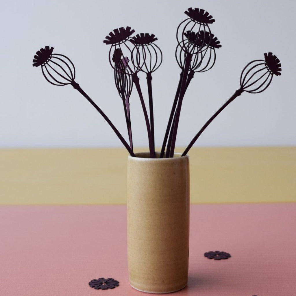 Dark purple, laser-cut poppy seed head decorations in a light brown cylindrical vase.