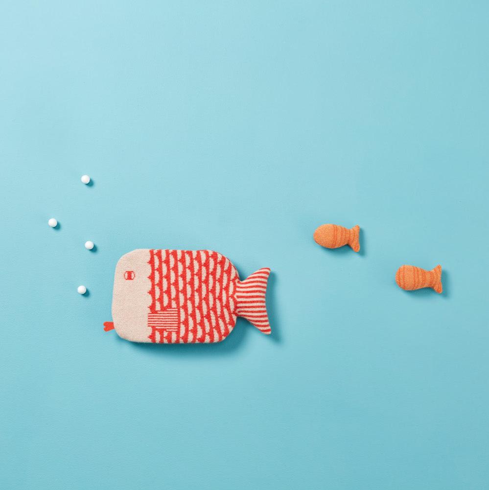 A red and cream fish hot water bottle by Donna Wilson swims against a light blue background with small white bubbles and orange fish.